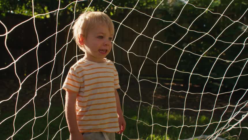 A happy toddler boy stands in a soccer goal in the backyard as the warm afternoon sun creates a beautiful lens flare