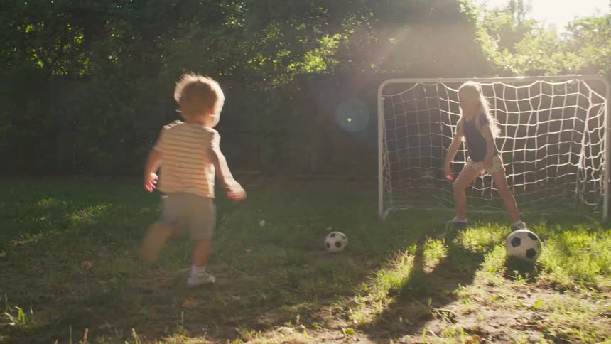 A toddler boy and his older sister play soccer together in their sun-drenched backyard