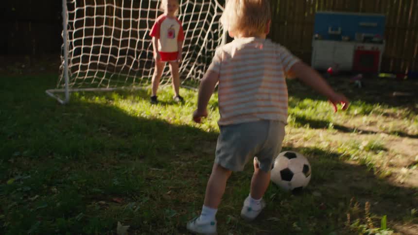 A happy little boy kicks a soccer ball while playing with his sister in their sunlit backyard