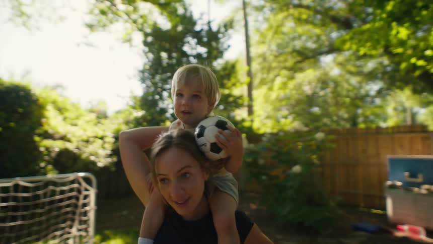 A joyful toddler holds a soccer ball while riding on his mother's shoulders as she walks through their sunlit backyard