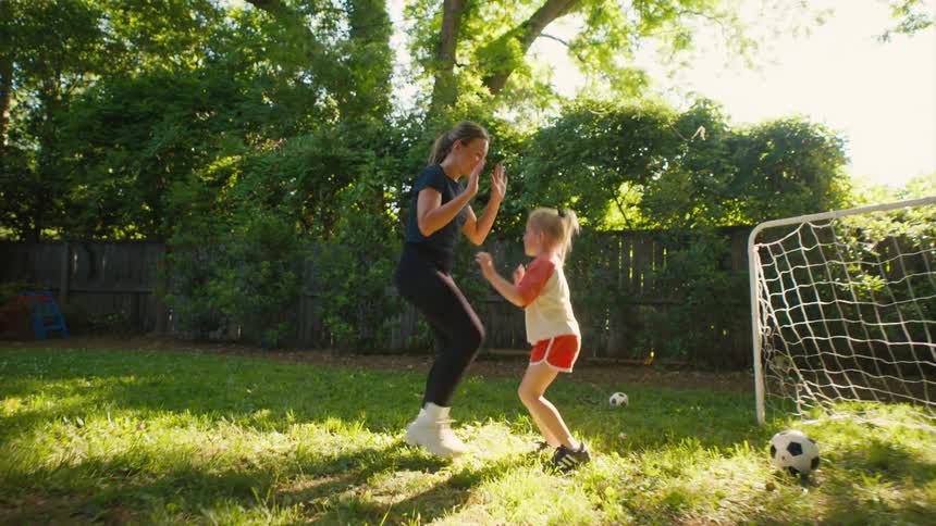 A mother and her young daughter playfully practice soccer drills in their sunny backyard, celebrating with a joyful high-five