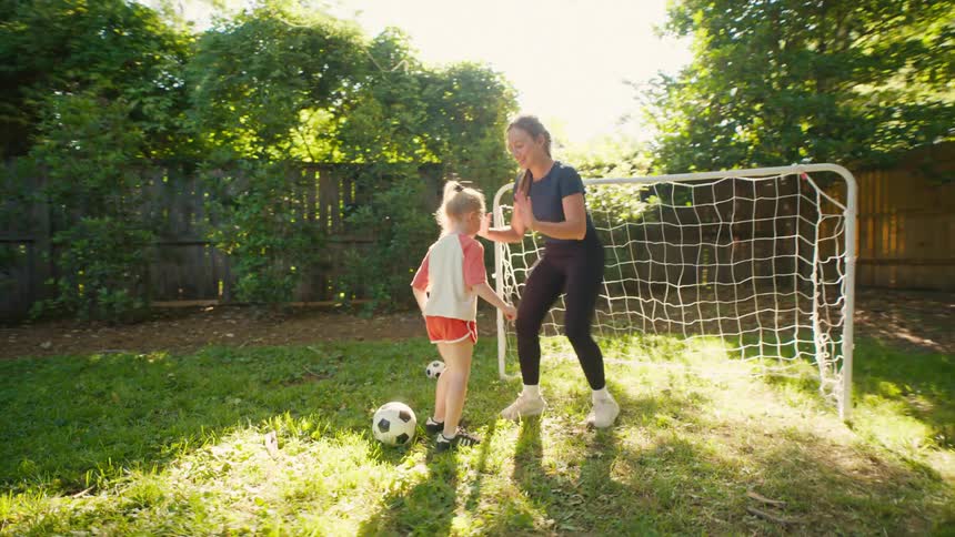 A proud mother and her young daughter celebrate with a joyful high-five while playing soccer in their sunny backyard