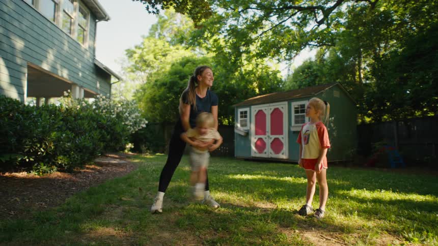 A mother playfully swings her young son in their sunny backyard while his sister watches