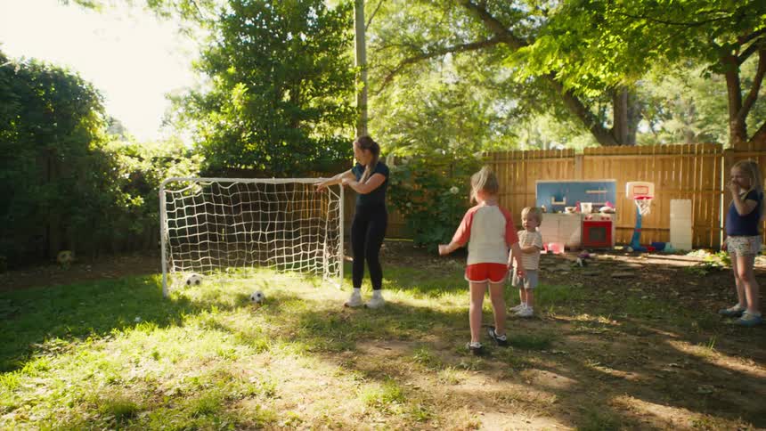 A woman teaches a group of young children how to head a soccer ball in their backyard on a sunny day