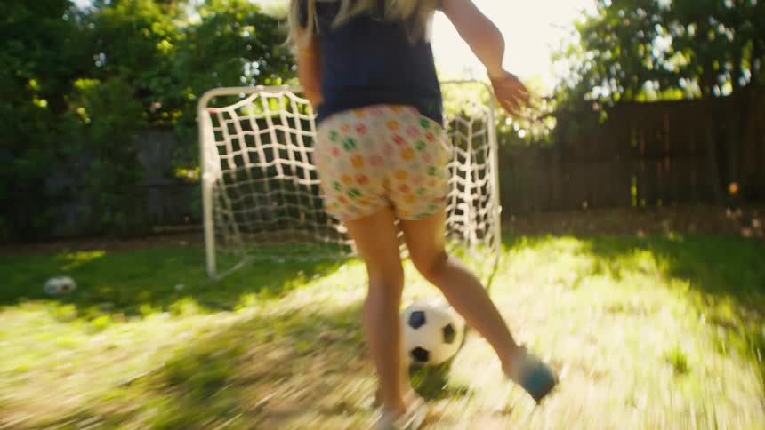 Low-angle slow-motion shot of a child scoring a soccer goal in a sunlit backyard