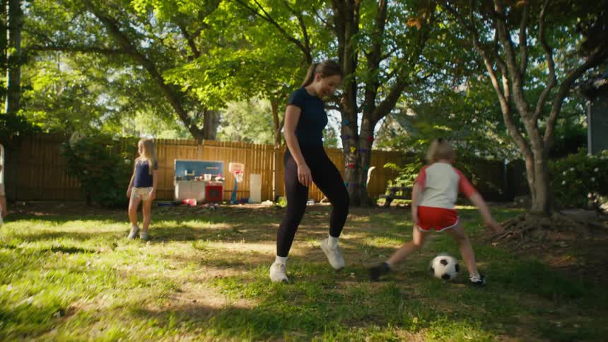 A young boy dribbles a soccer ball in the backyard while his mother playfully tries to take it