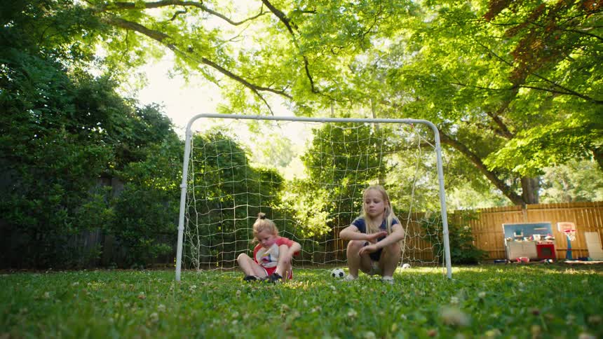 Two young sisters run to their soccer goal and sit down for a break in their lush, green backyard