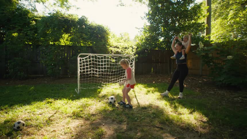 A young girl scores a soccer goal in the backyard while her mom, holding her toddler sister, cheers her on