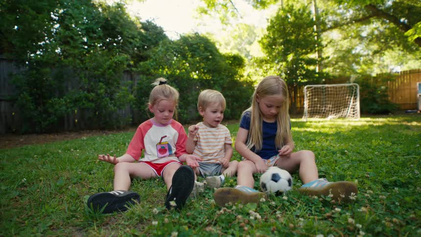 Three young siblings play together with a small soccer ball while sitting in the grass of their backyard on a sunny day