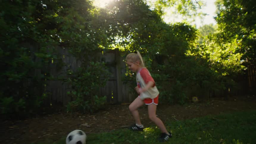 Slow-motion shot of a happy young girl scoring a soccer goal and dribbling the ball in her sunny backyard