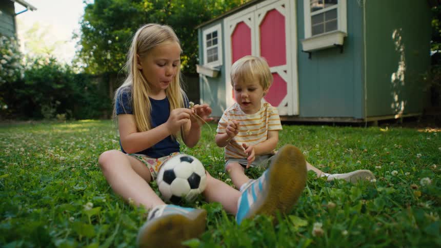 A young girl and a toddler boy sit together in the lush green grass of their backyard, playing with clovers on a sunny day