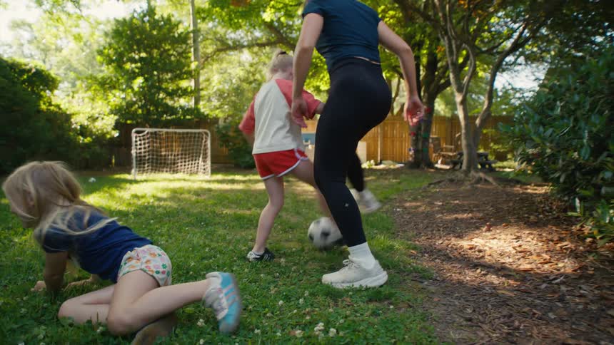 A mother and her two young daughters playfully wrestle for a soccer ball before running around and playing in their sun-drenched backyard