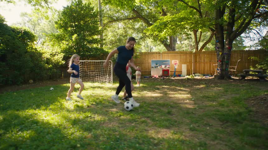 A mother and her young children have fun playing a game of soccer in their sunny backyard