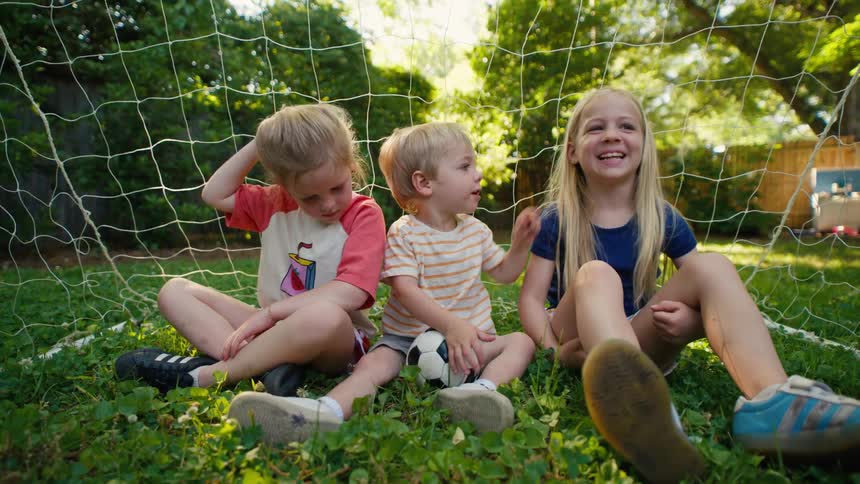 Playful siblings, two girls and a toddler boy, share a sweet moment while sitting in a soccer goal in their backyard