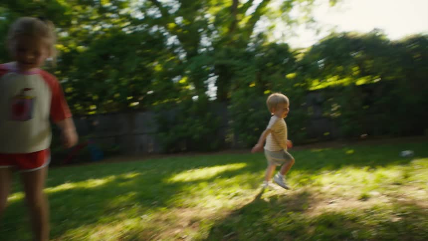 A mother dribbles a soccer ball in the backyard while her laughing daughter and toddler son playfully chase after her
