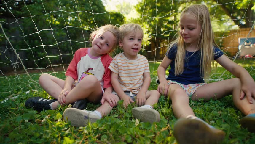 Three young siblings, two sisters and a little brother, sit together in a soccer goal in their backyard on a sunny summer day