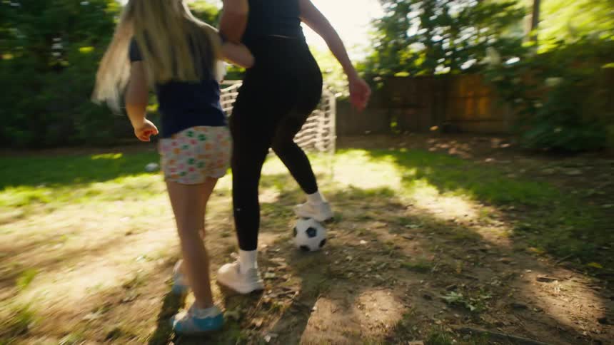 An athletic mother teaches her young daughter soccer skills in their sunny backyard before the girl kicks the ball towards the net