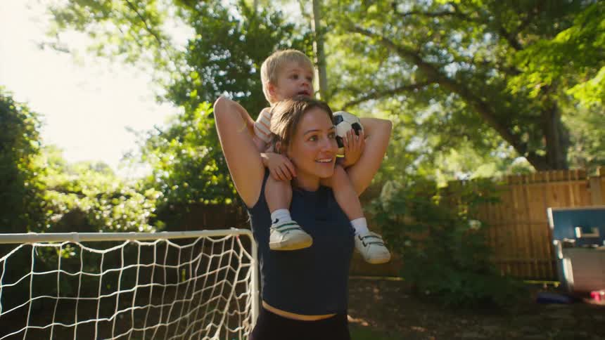 A happy mother carries her toddler son on her shoulders in their sunny backyard with a soccer goal