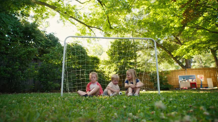 Three young siblings sit together inside a soccer goal in their green backyard, taking a break from playing on a sunny day