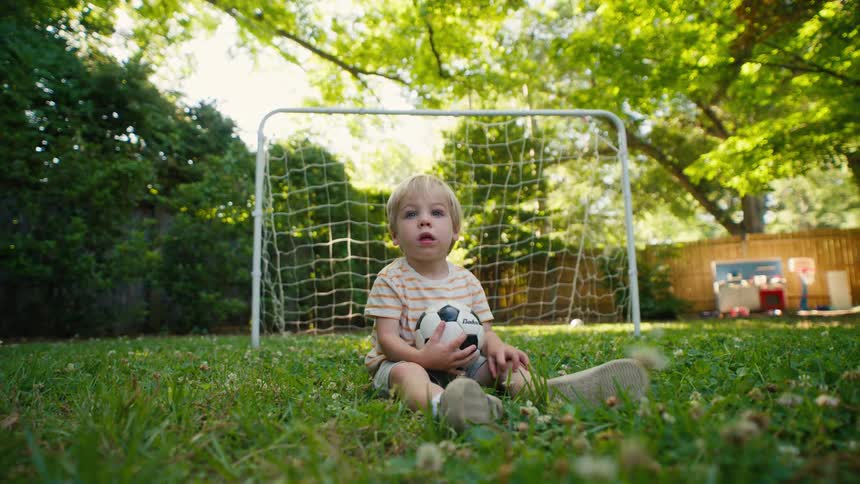 A toddler boy being handed a soccer ball by his sister before standing up and dropping it in the backyard