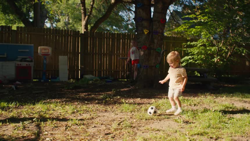 A young boy runs with a soccer ball and kicks it towards his sister playing goalie in their sunny backyard