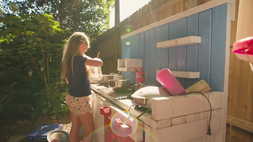 An imaginative young girl with long blonde hair pretends to cook at her outdoor mud kitchen on a sunny summer day