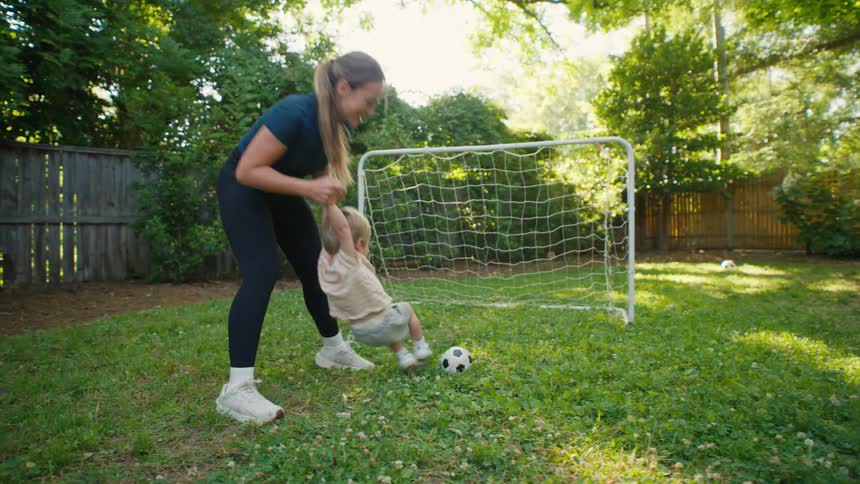A smiling mother swings her toddler by his arms as he kicks a soccer ball towards the goal in their backyard