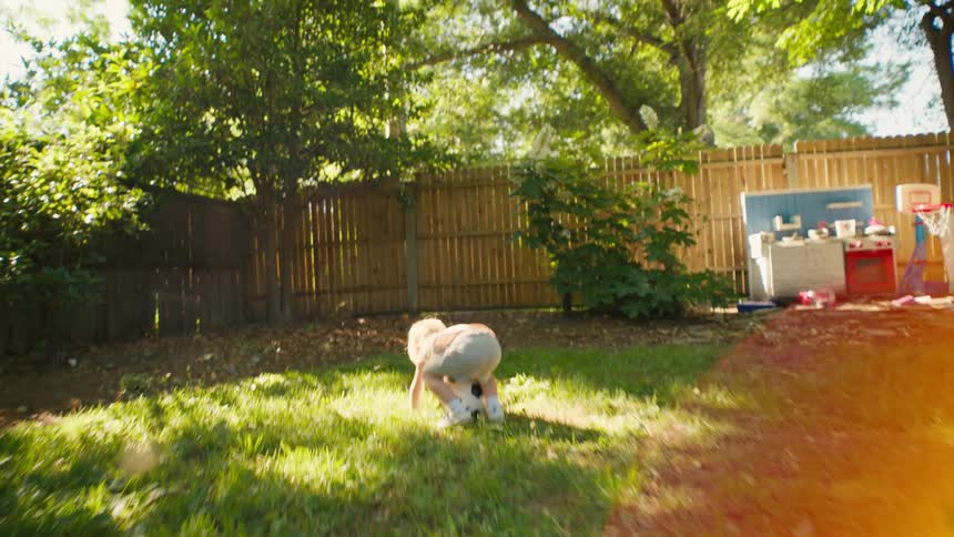 A determined little boy falls down while running across a sunny backyard but gets right back up to joyfully grab his soccer ball