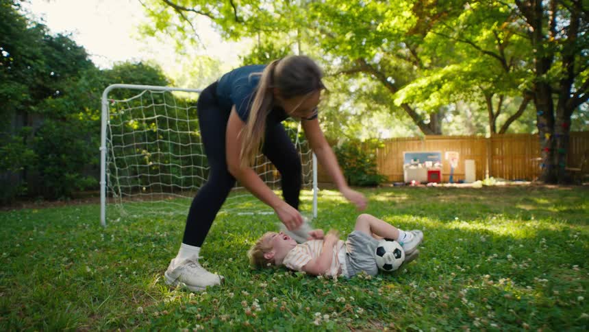 A mother and her toddler have a playful soccer game in the backyard which ends in a tickle fight