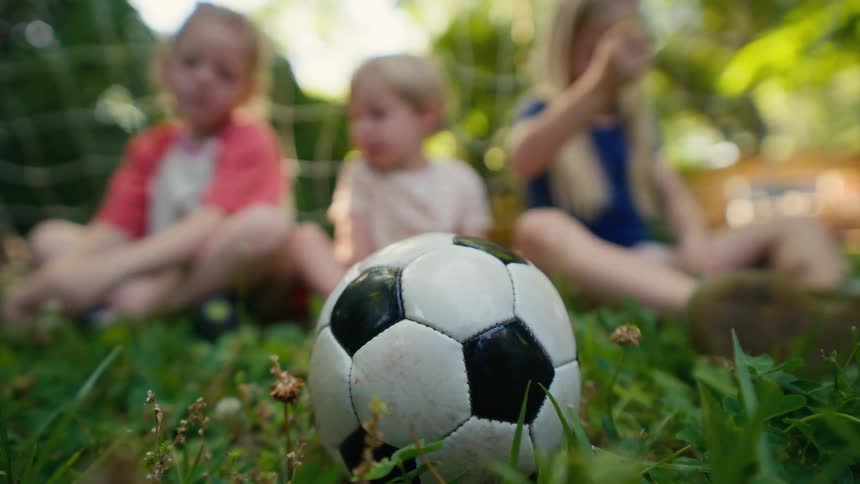 A focus pull from a soccer ball on the grass reveals three young siblings sitting and relaxing together in their backyard