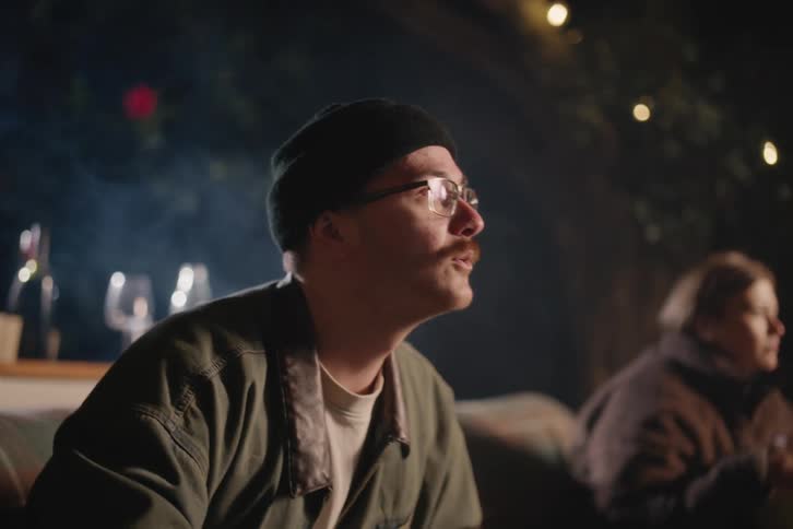 A young man in a beanie and glasses smiles as he watches a movie with friends during a cozy backyard gathering at night