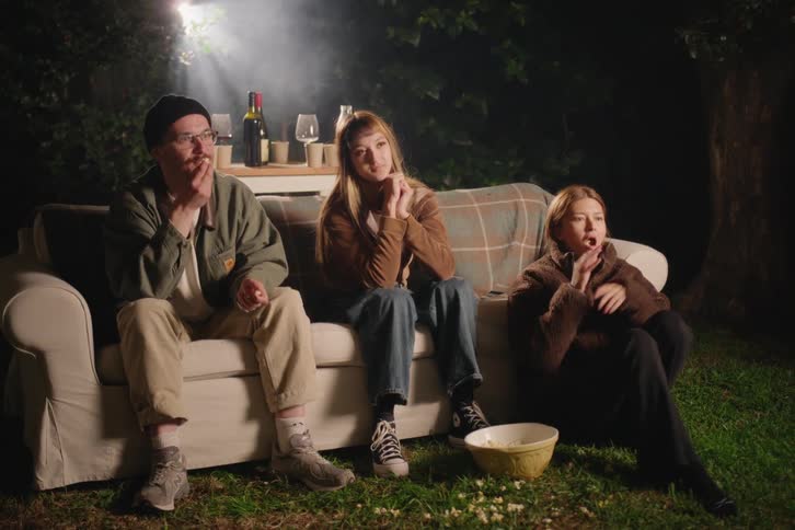 Three friends enjoy an outdoor movie night in the garden, sitting on a sofa and eating popcorn while watching a film