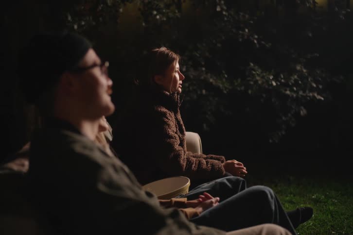 A young couple sits in their backyard at night, captivated by the glow of an outdoor movie screening