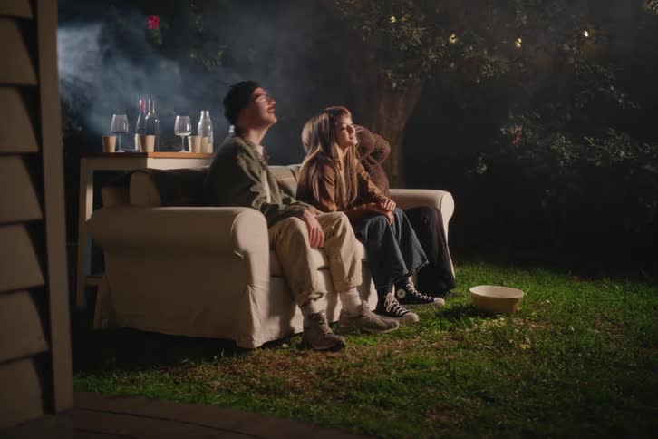Three friends react with fright and laughter while watching a movie on a projector in their backyard at night