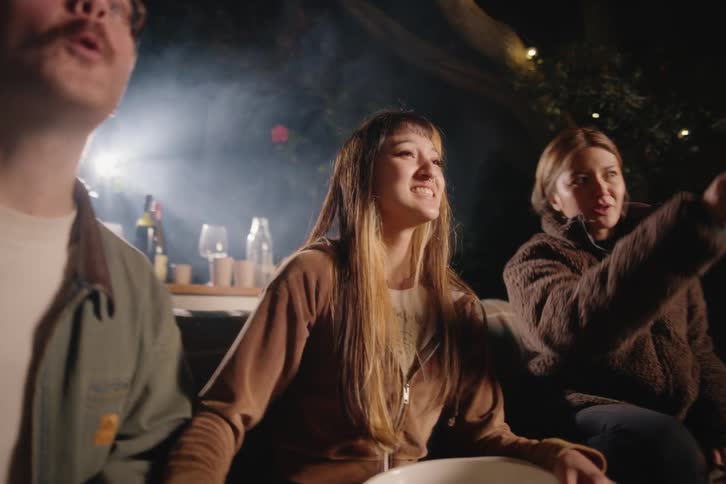 Three excited friends watch an outdoor movie on a projector at night, sitting together on a couch in the backyard