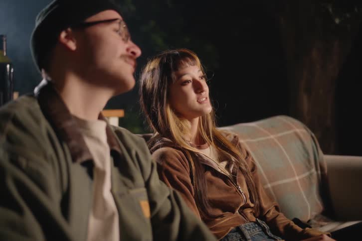 A young woman talks with a friend as they sit on a couch during a cozy backyard hangout at night