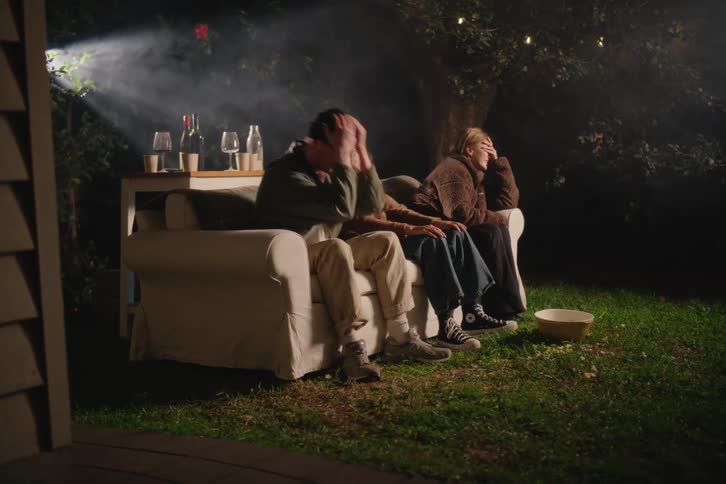 A group of friends on a couch in the backyard at night react with shock and laughter while watching a movie on a projector