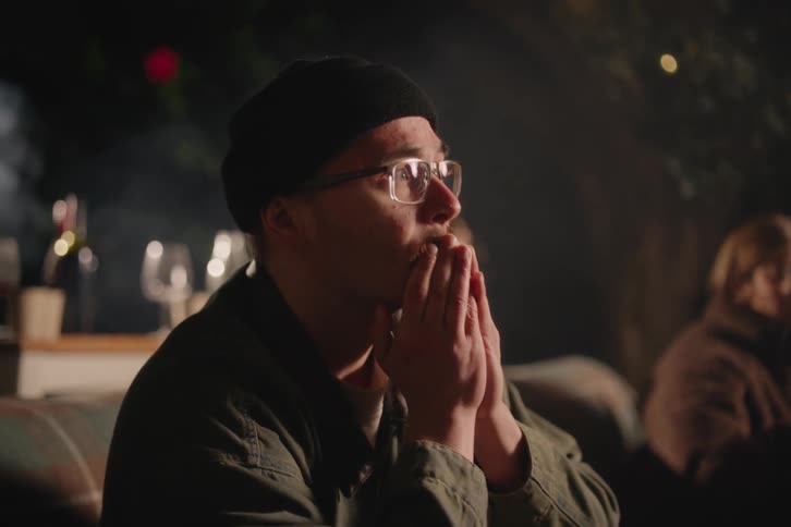 A young man with glasses and a beanie anxiously watches a movie with friends during an outdoor screening at night
