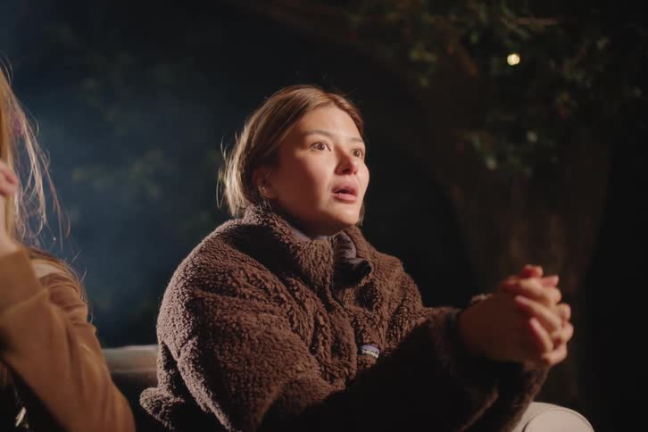 A young woman's expressive face shows a range of emotions as she listens and speaks during an outdoor nighttime gathering