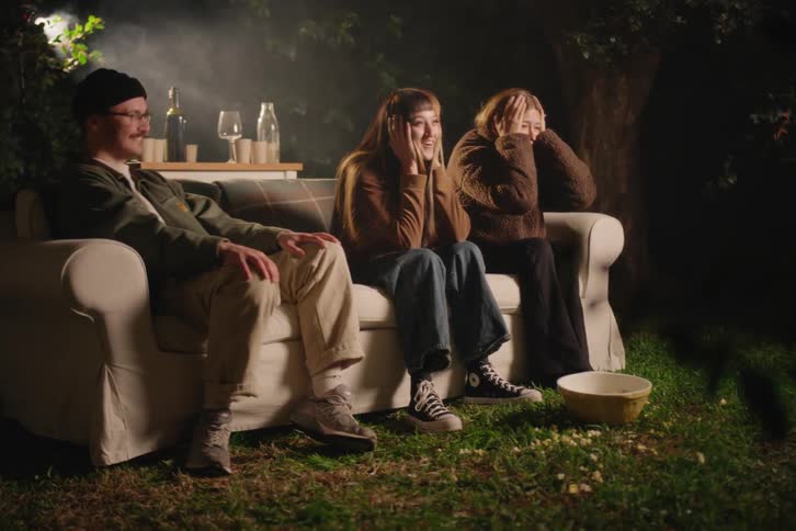 A group of friends have an outdoor movie night, reacting with fear and laughter while watching a film on a projector in their backyard