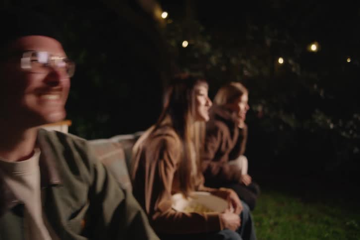 A group of young friends sit on a couch for a cozy backyard movie night, eating popcorn and watching a projector screen