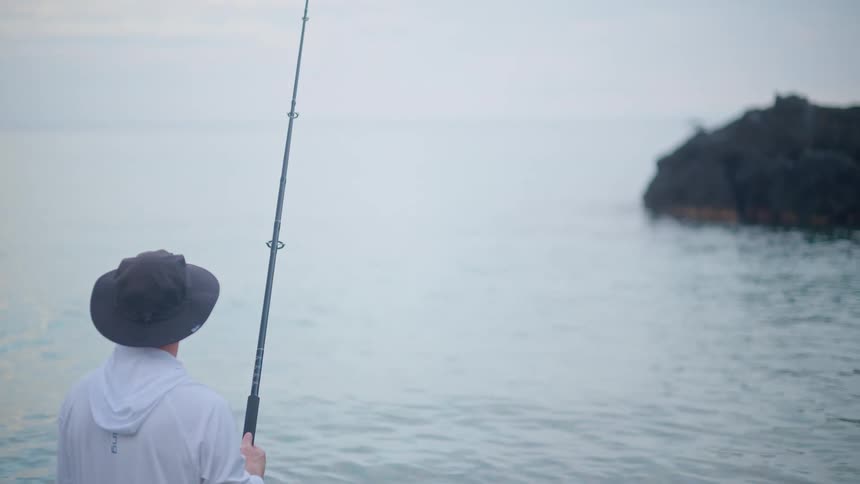 A lone fisherman in a hat stands in the calm water at dawn, patiently waiting for a bite