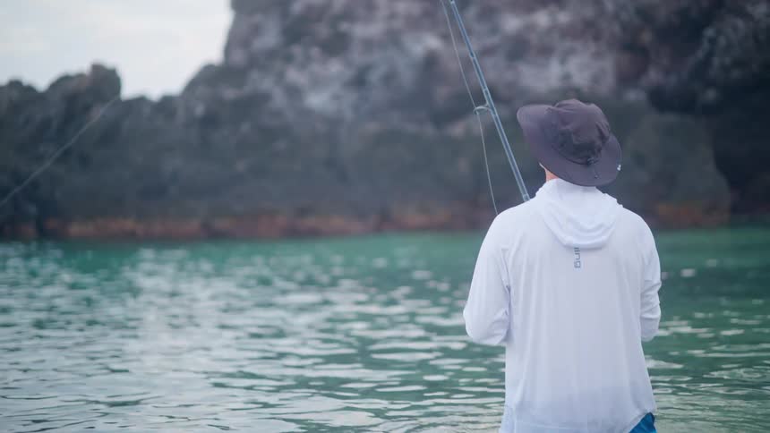 A man wearing a hat stands in the ocean with a fishing pole near a rocky coastline