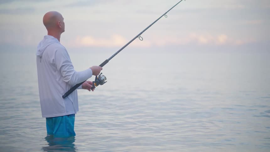 A man stands patiently in the calm ocean water while fishing during a peaceful, hazy morning