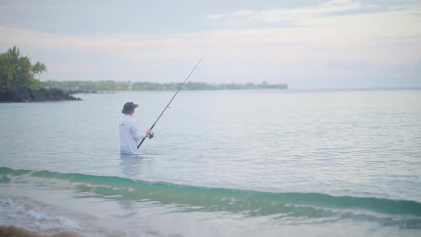 A lone fisherman stands patiently in the calm, shallow ocean water during a peaceful, hazy morning