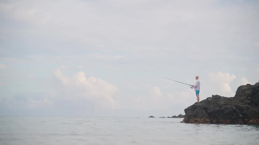 A lone fisherman stands on a rock formation in the calm ocean, enjoying a peaceful morning of fishing under a cloudy sky