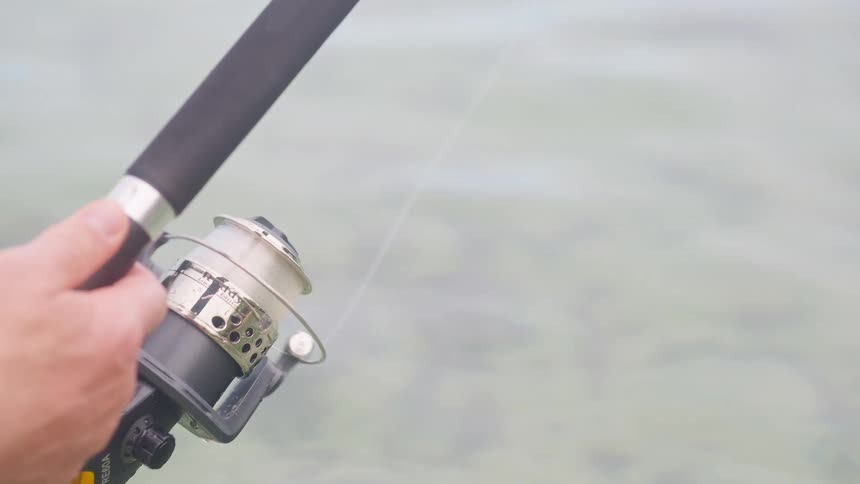 A close-up shot of a fisherman reeling in the fishing line on a spinning rod over calm water