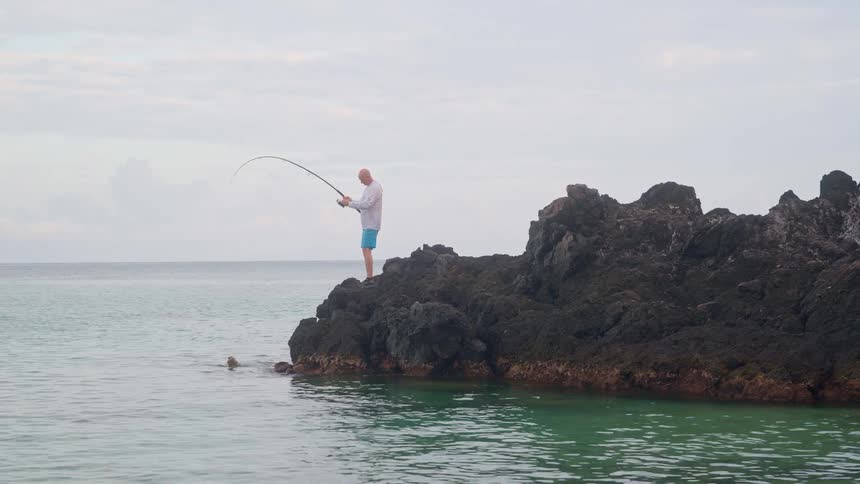 A man standing on a large rock in the ocean reels in a fish he has just caught on his line