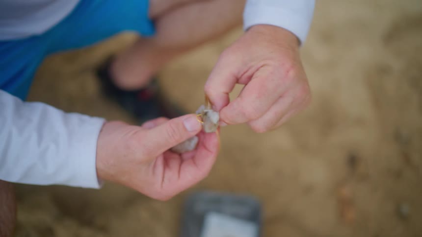 A fisherman carefully baits a hook with a shrimp while preparing to go fishing on the sand