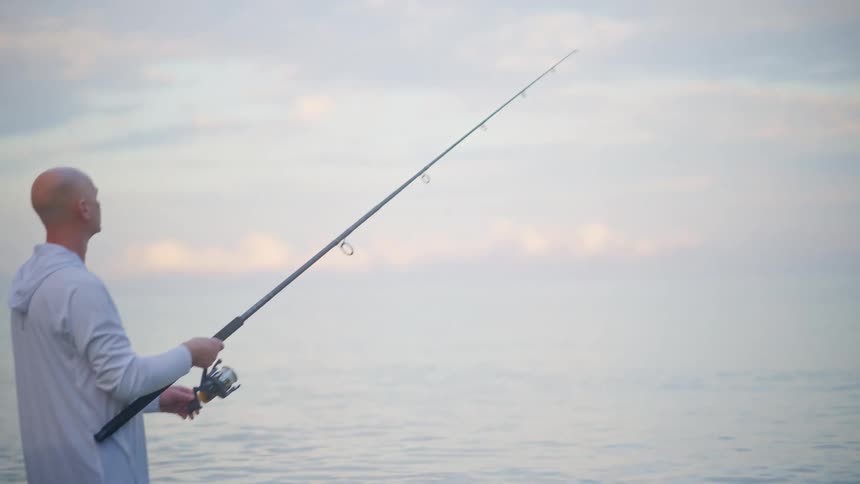 A slow reveal shot pans down to a solitary man fishing in the calm ocean at dawn
